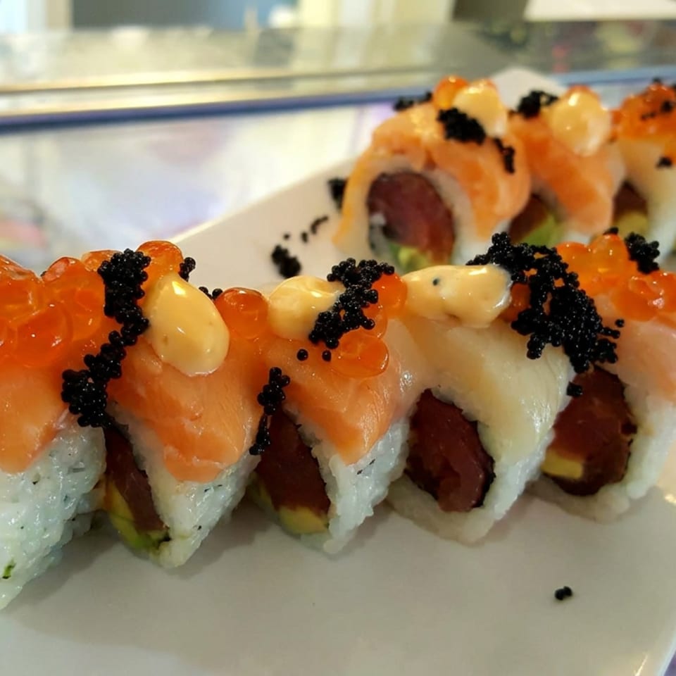Close-up of sushi rolls topped with salmon, orange roe, black tobiko, and creamy sauce on a white plate.