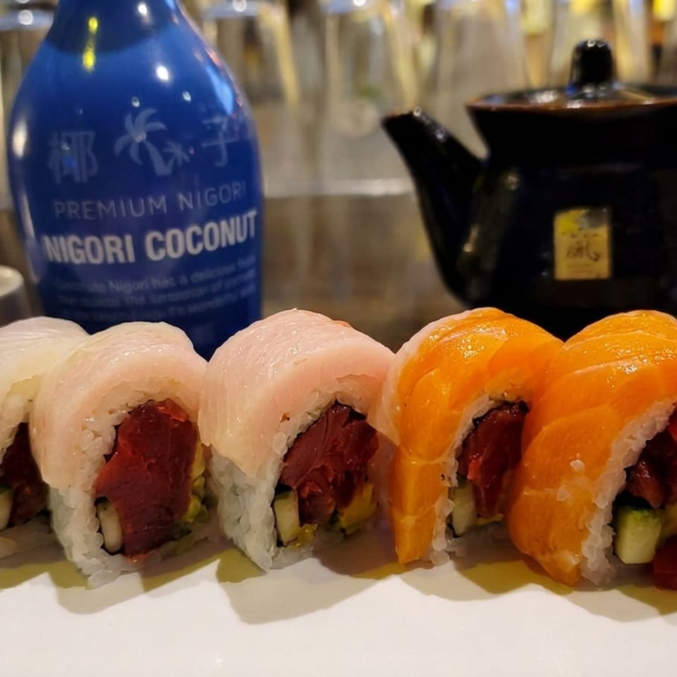 Colorful sushi rolls arranged on a white plate with nigori coconut sake and a black teapot in the background.