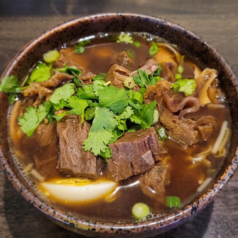 Bowl of dark broth with tender beef, noodles, cilantro, green onions, and a halved boiled egg on a wooden table.