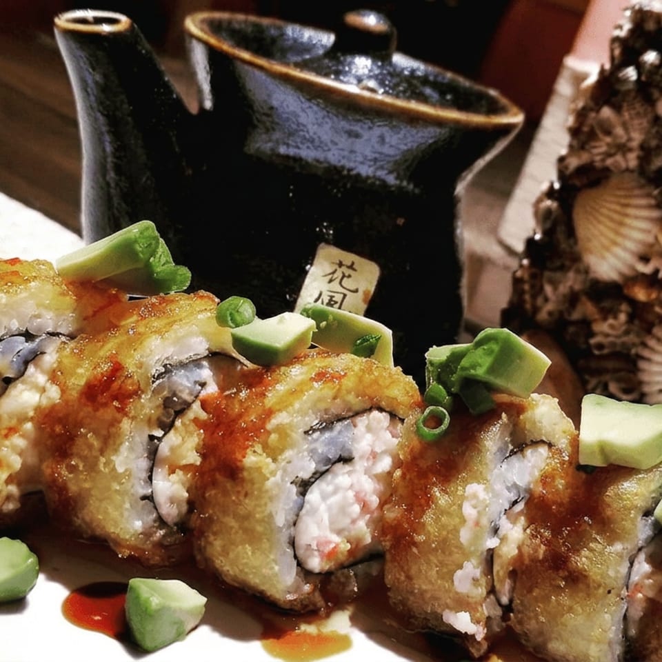 Tempura sushi roll topped with avocado and green onions, served with dipping sauce beside a shell-covered decorative centerpiece.