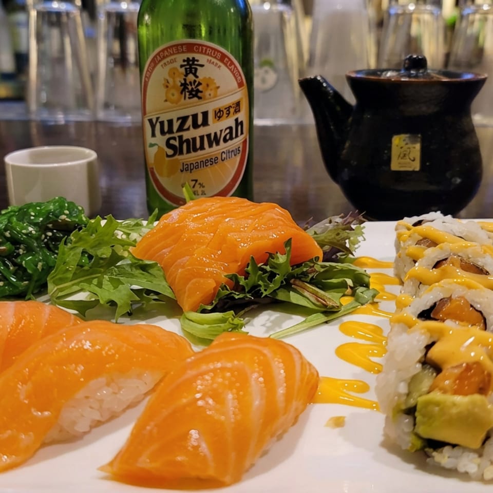 Plate of salmon sushi, sushi rolls with orange sauce, seaweed salad, and greens beside yuzu shuwah and a black teapot.