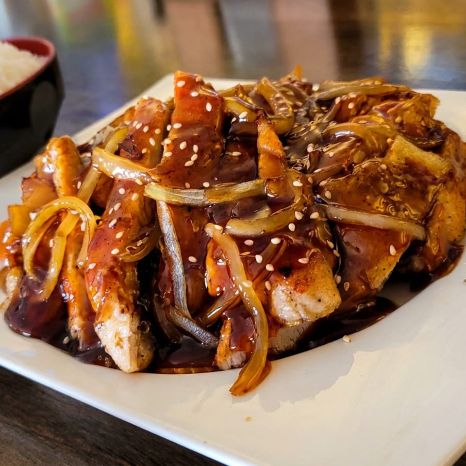 Glazed stir-fried chicken with onions and vegetables, sprinkled with sesame seeds, served alongside a bowl of white rice.