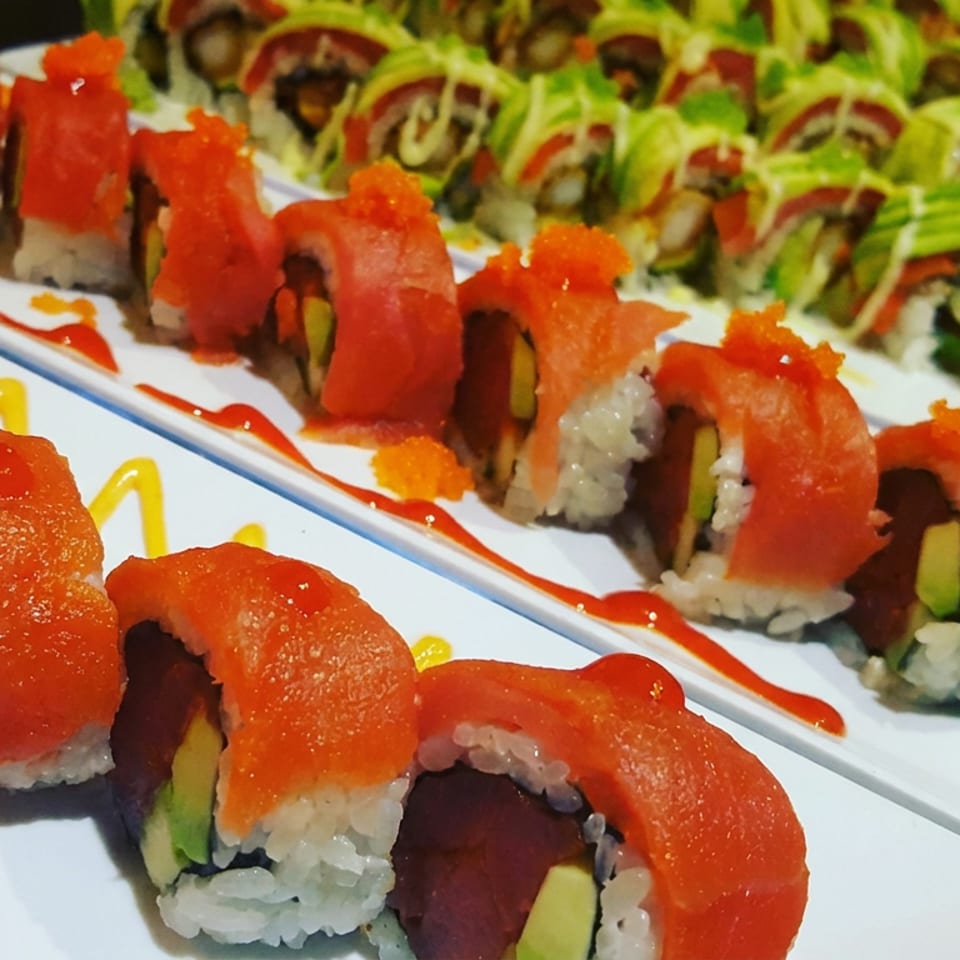 Plates of salmon-topped sushi rolls with avocado, rice, colorful sauces, and shredded lettuce garnish.