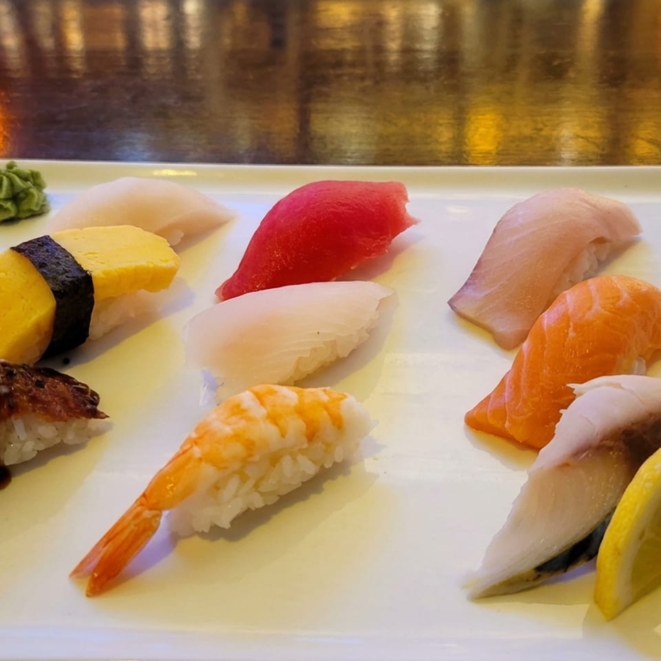 Assorted sushi, including shrimp, tuna, salmon, squid, eel, and tamago, arranged on a white plate with wasabi, lemon, and ginger.
