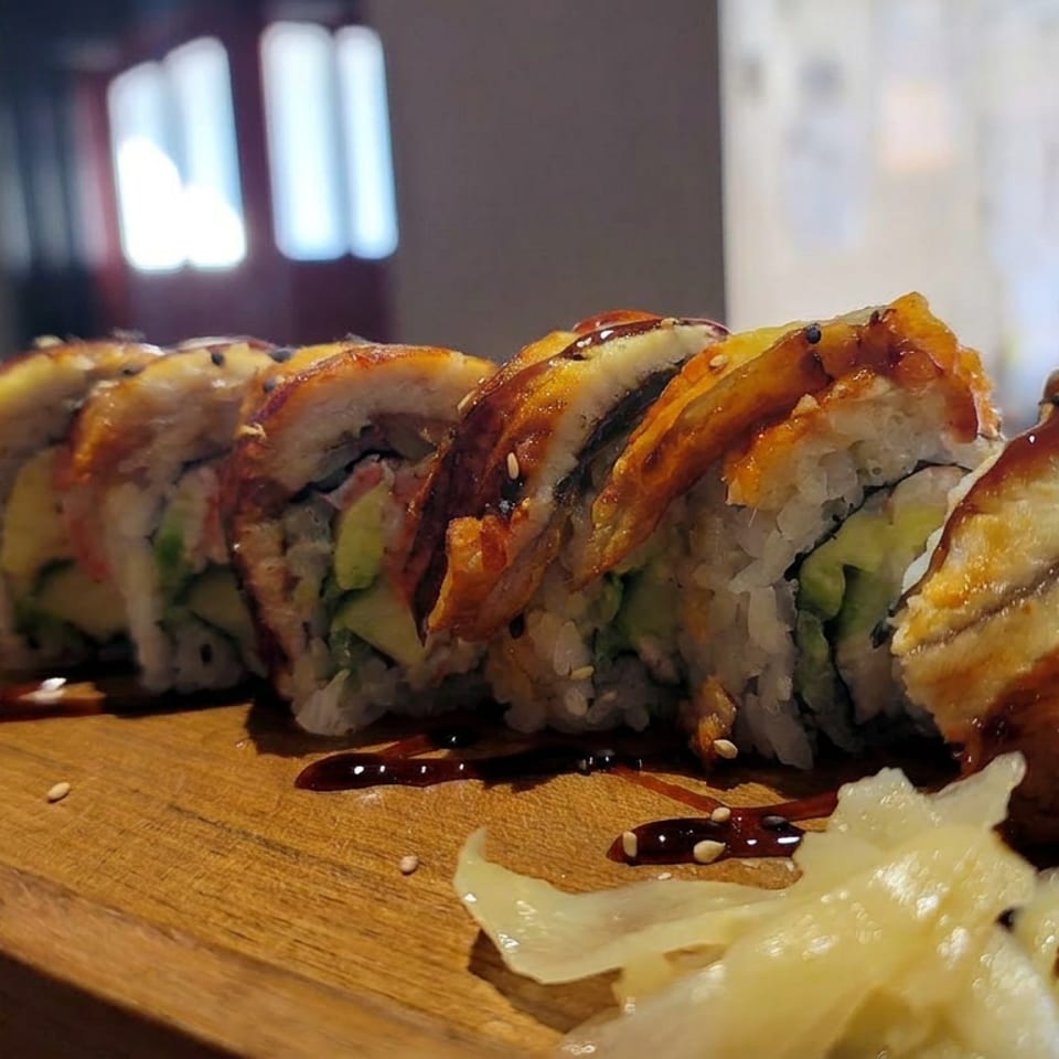 Glazed sushi rolls topped with grilled fish and vegetables, arranged on a wooden serving board with sesame seeds and sauce.