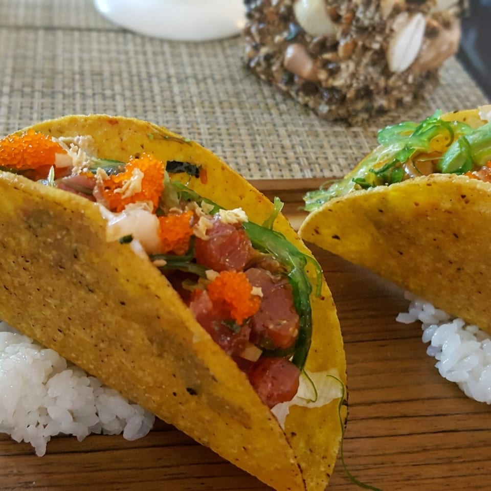 Four crispy taco shells filled with sushi rice, diced raw fish, orange roe, greens, and shredded toppings on a wooden board.