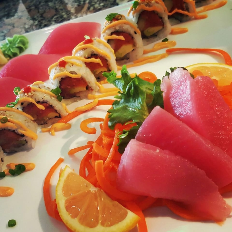 Sushi roll slices drizzled with orange sauce alongside pink sashimi, shredded carrots, citrus, and greens on a white plate.