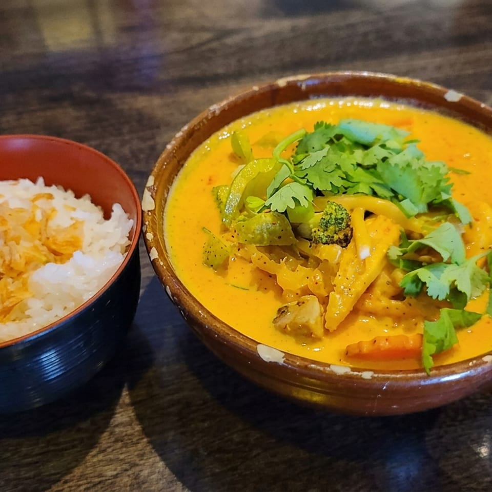 Bowl of creamy yellow curry with vegetables and cilantro beside white rice on a dark wooden table.