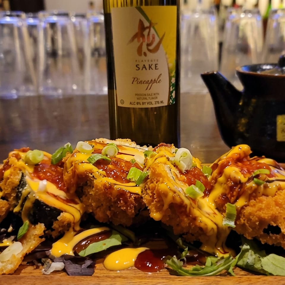 Crispy sushi rolls drizzled with sauces and scallions, served with greens and ginger beside pineapple sake and a black teapot.